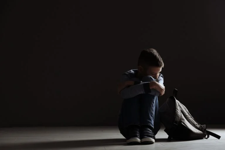 Upset boy with backpack sitting in dark room.