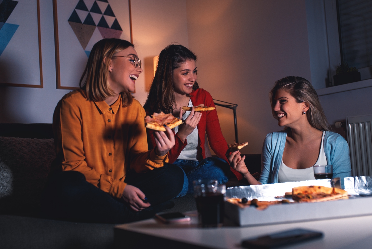 Three female friends chatting and enjoying eating pizza at home.