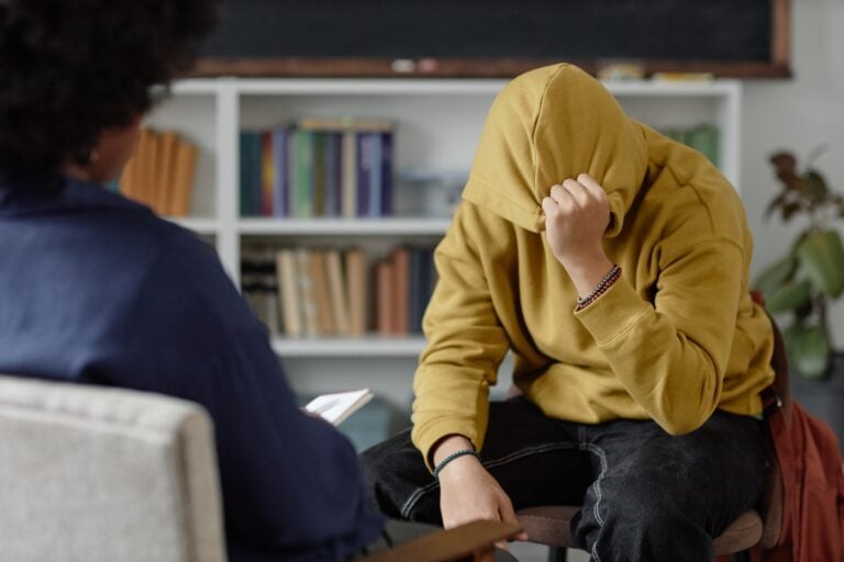 distressed teenage boy sharing mental health troubles with therapist while sitting on chair and hiding face during one on one session