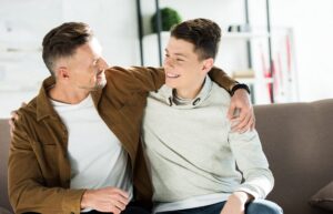 smiling father and teen son hugging on sofa at home and looking at each other