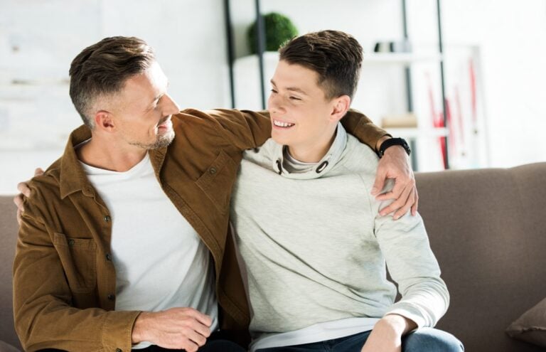 smiling father and teen son hugging on sofa at home and looking at each other