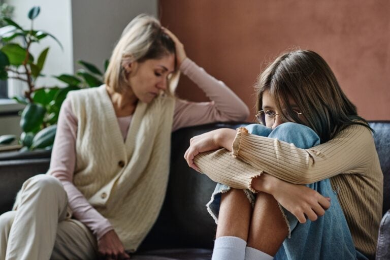 upset teenager sitting on a sofa with image of a distressed parent in the background